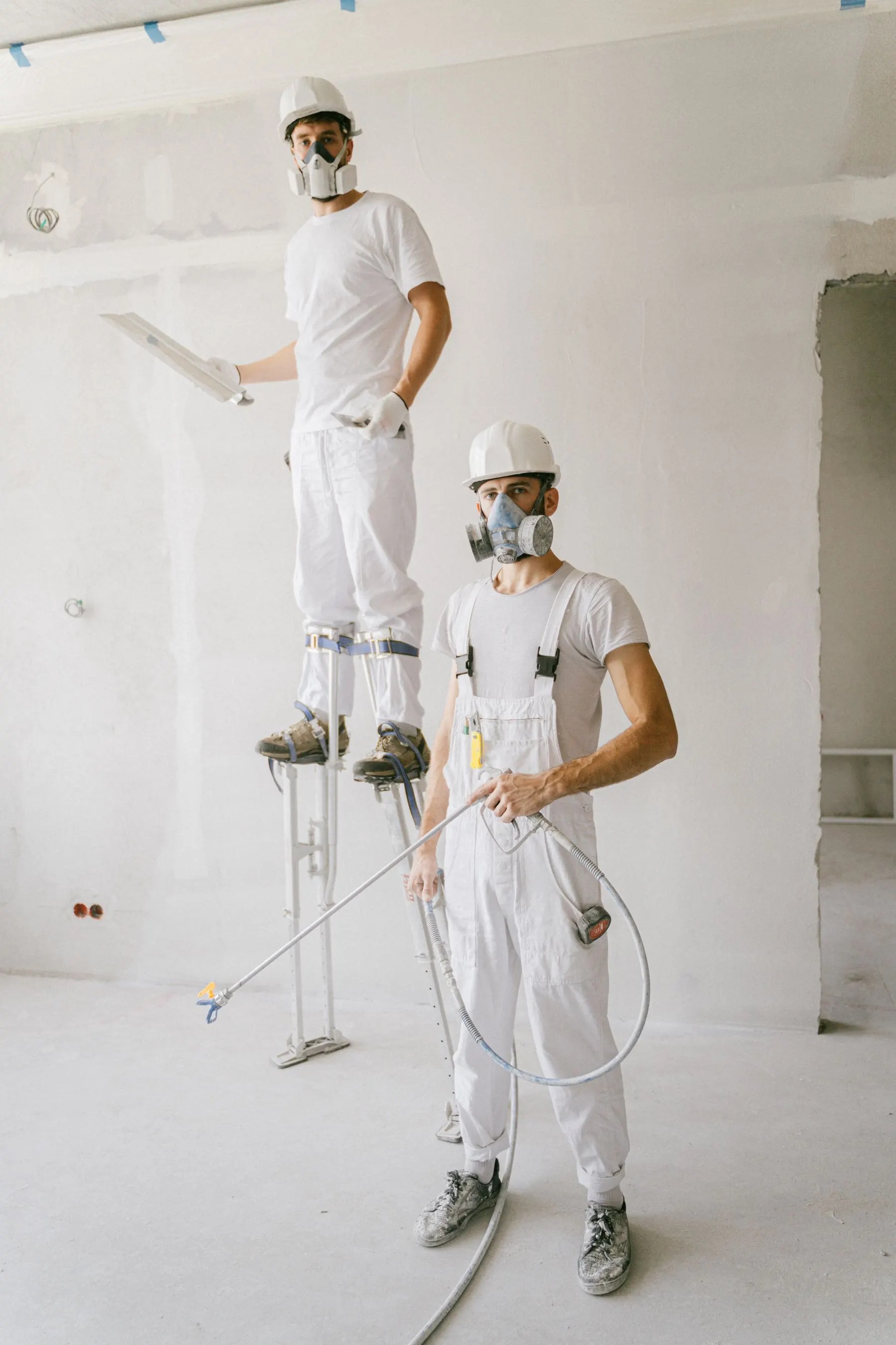 Two painters in white overalls and respirators preparing a room, one working from stilts